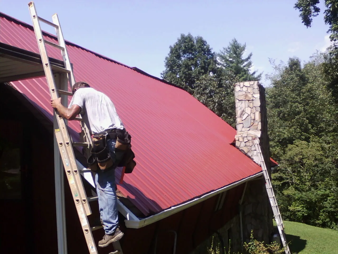 Roofer on a ladder beside a red metal roof and stone chimney — Soffit & Fascia Repair by Roofing Brothers in Deweyville